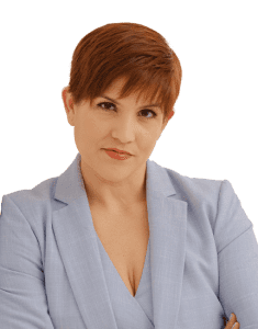 A person with short reddish-brown hair, wearing a light blue blazer, looks at the camera with a neutral expression and arms slightly crossed.
