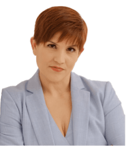 A woman with short reddish-brown hair wearing a light blue blazer, looking at the camera with a neutral expression against a plain background.