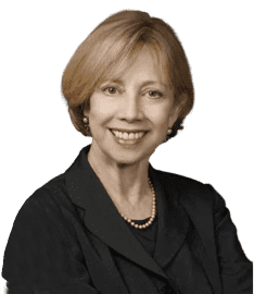 A woman with short, light brown hair wearing a black blazer, pearl necklace, and smiling against a plain light background.
